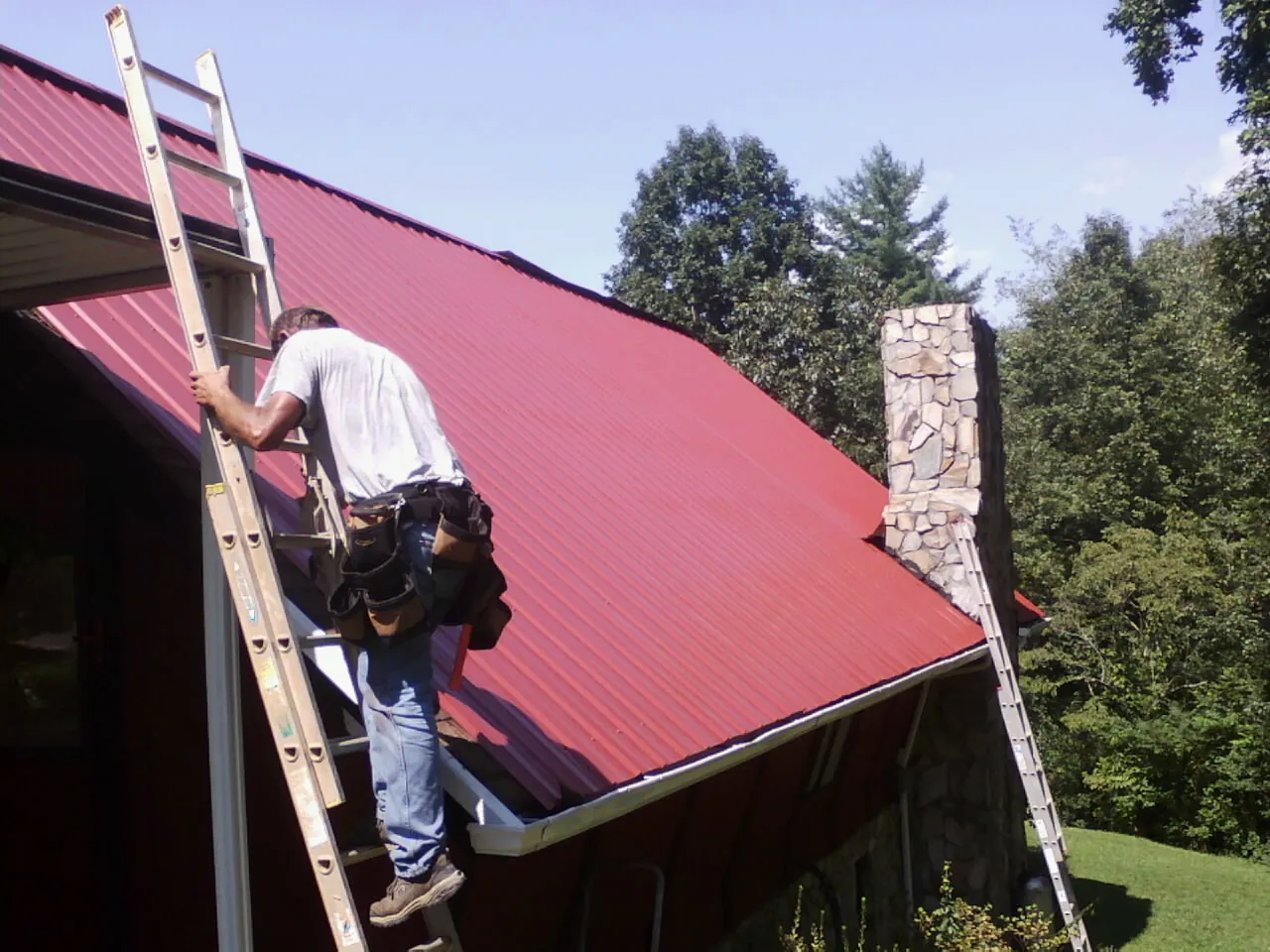 Roofer on a ladder beside a red metal roof and stone chimney — Soffit & Fascia Repair by Roofing Brothers in Collierville